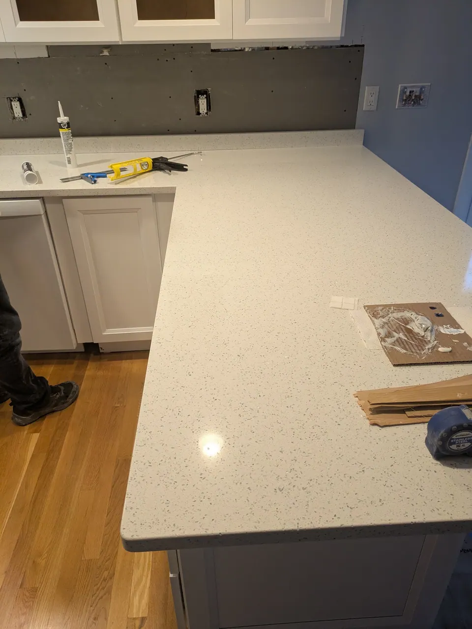In progress: speckled white peninsula countertop with a caulk gun and tools, bare wall behind.