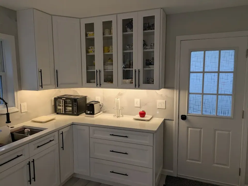 Glass-front and solid white wall cabinets over a lit counter with drawer bases, next to a white door.