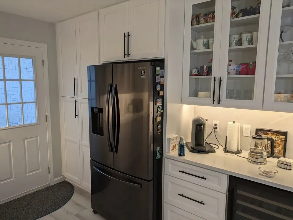 Tall white pantry cabinets beside a dark refrigerator, with glass-front cabinets and drawers to the right.