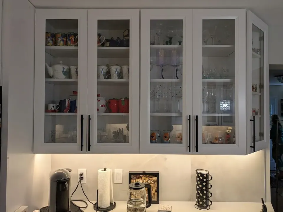 Glass-front wall cabinets holding mugs and glassware, with under-cabinet lighting over the coffee counter.