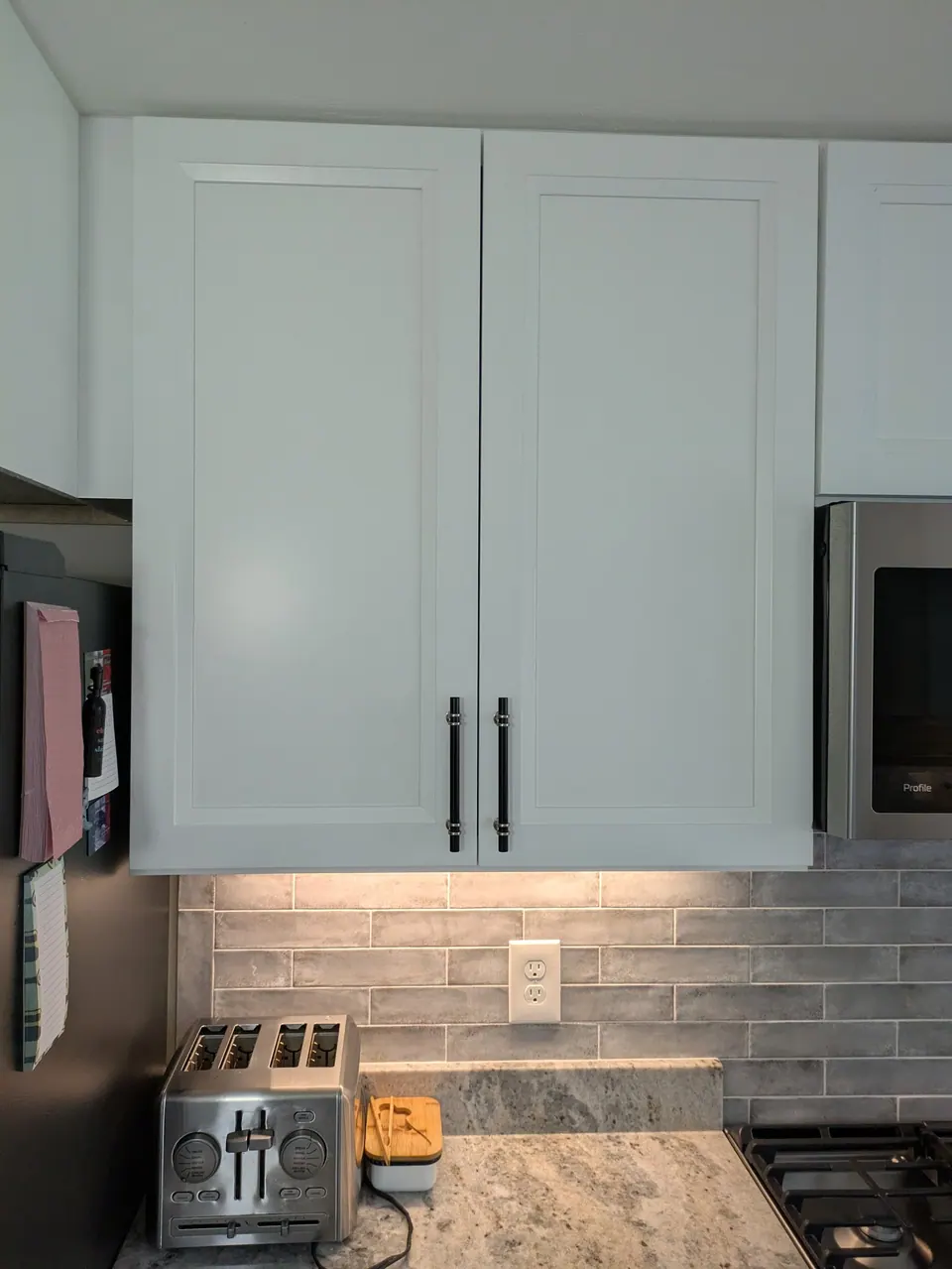 White two-door wall cabinet with black bar pulls above a gray tile backsplash.