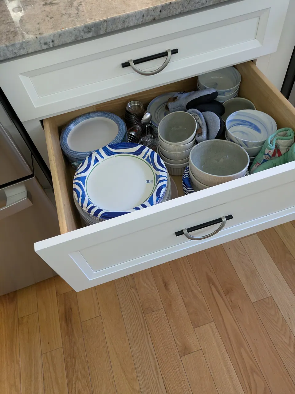 White deep drawer open with stacked plates and bowls, wood-look floor below.