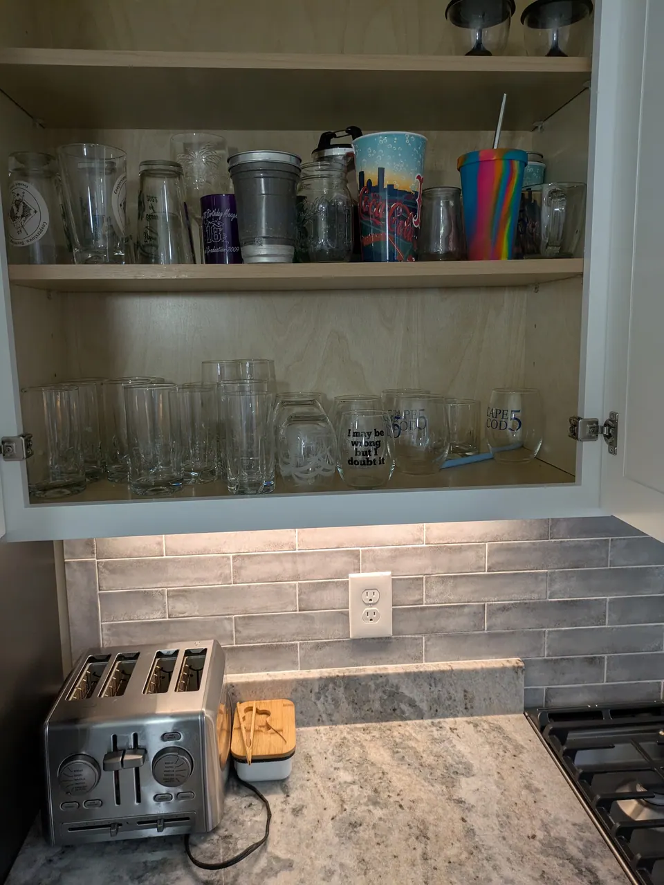 White wall cabinet open, showing two shelves of drinking glasses above the countertop.