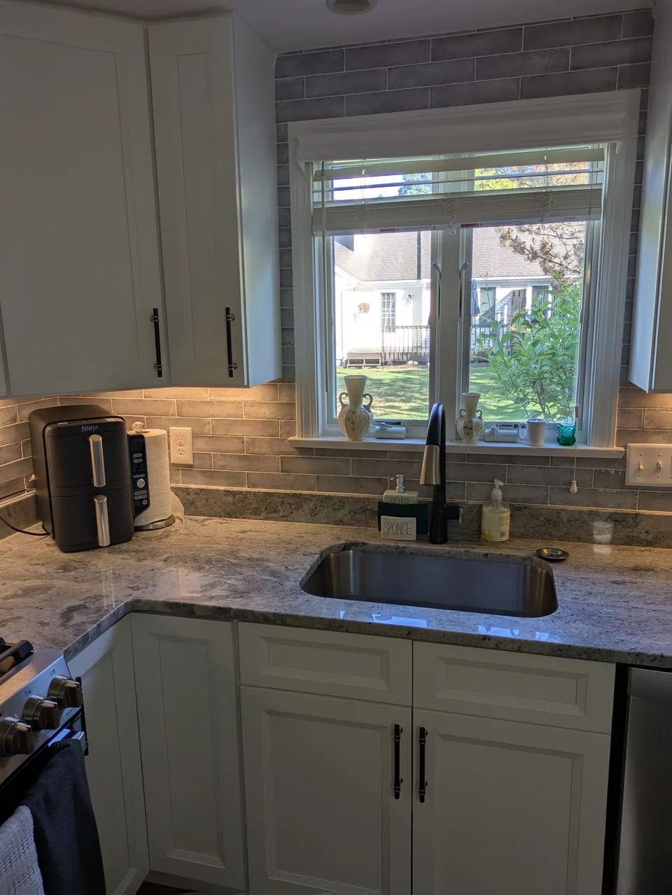 White sink base and wall cabinets around a window, black faucet, gray tile backsplash.