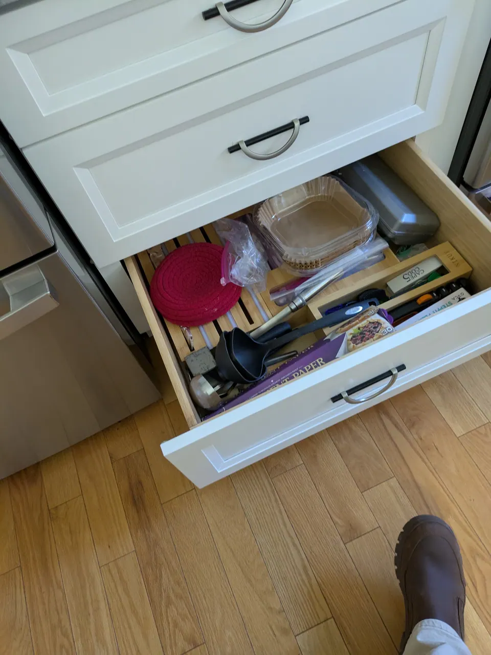 White bottom drawer open with organizer trays and utensils, wood-look floor below.