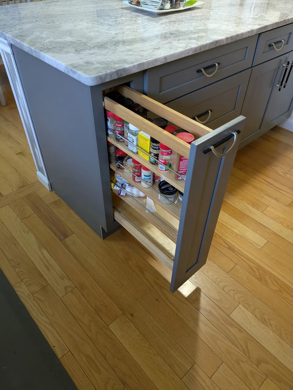 Gray peninsula end cabinet with a pull-out spice rack open, jars on wood shelves.