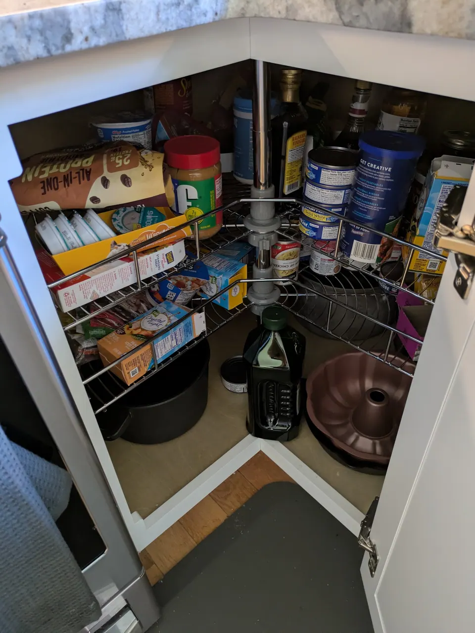 White corner base cabinet open, showing a two-tier wire lazy Susan with groceries.
