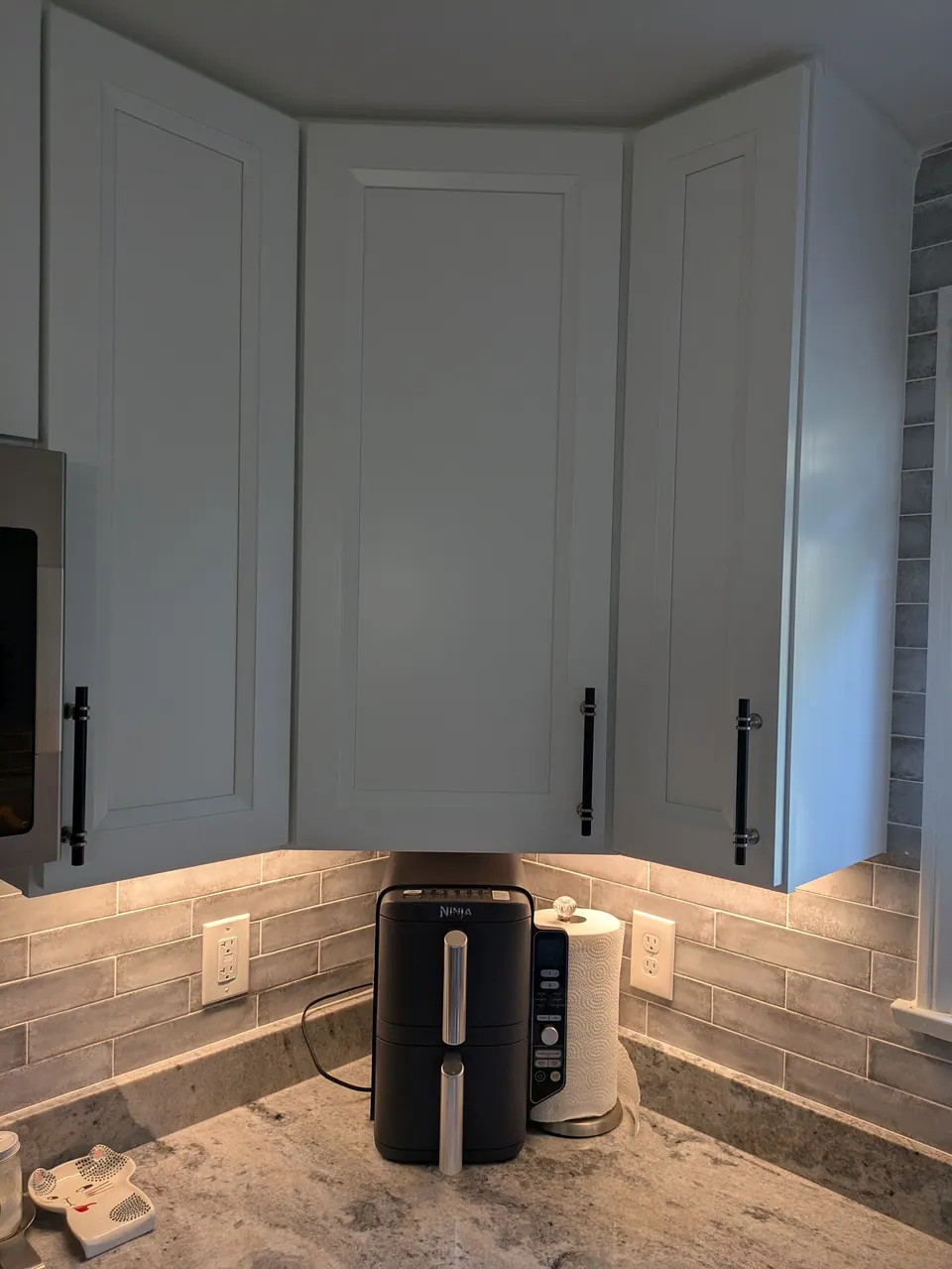 White corner wall cabinets with black bar pulls and under-cabinet lighting on gray tile.