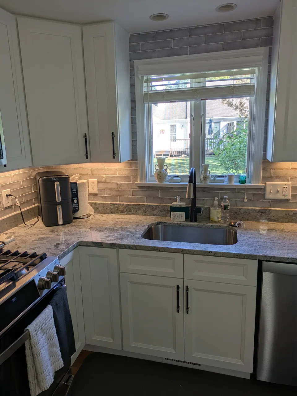 Sink wall of white shaker cabinets with a black faucet under a window and a gray tile backsplash.