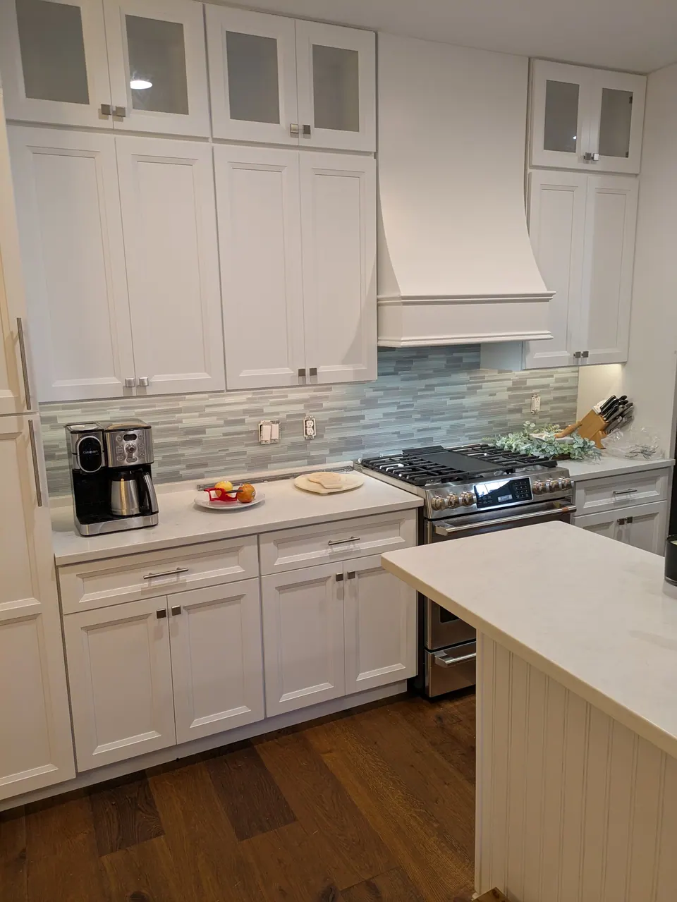 White kitchen with lit glass-top wall cabinets, a white wood range hood and an island with a white quartz top.