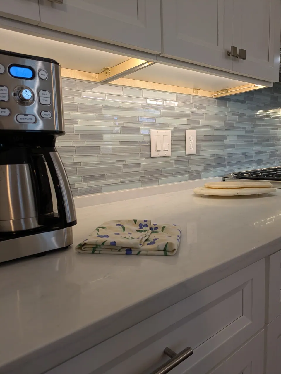White quartz counter under lit wall cabinets with a glass mosaic backsplash.