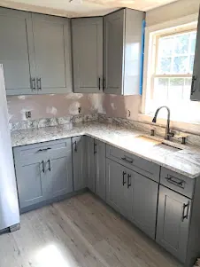 Gray shaker wall and base cabinets in a corner with speckled countertop and sink under a window.