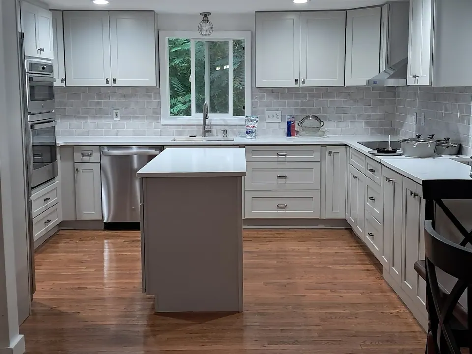 U-shaped kitchen with light gray shaker cabinets, small island, wall ovens and sink under a window.